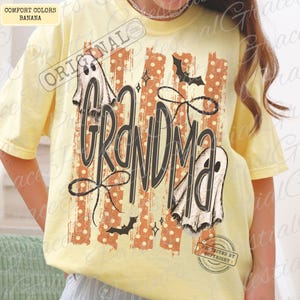 May include: A pale yellow short-sleeve t-shirt featuring a graphic print with the word GRANDMA in large black script. The design includes two white ghosts, black bats, and a background of vertical orange stripes with white polka dots.