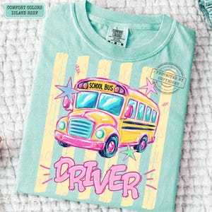 May include: A light teal t-shirt featuring a yellow school bus graphic with the text "SCHOOL BUS" above the windshield and "DRIVER" in large pink letters below. The design includes light yellow vertical stripes and pink and blue star accents.