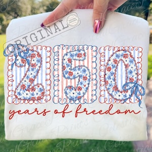 以下が含まれることがあります： 白いTシャツ。数字の「250」は花柄で、赤と青で縁取られ、「2」と「0」には青いリボンが付いています。「years of freedom」の文字が赤色の筆記体で下に書かれています。