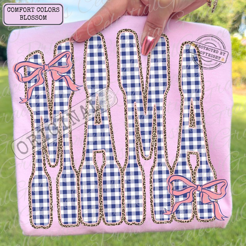 May include: A pale pink t-shirt featuring the word "MAMA" in a blue and white gingham design, edged with leopard print. Pink bows adorn the top left and bottom right. The text "COMFORT COLORS BLOSSOM" is in the top left.