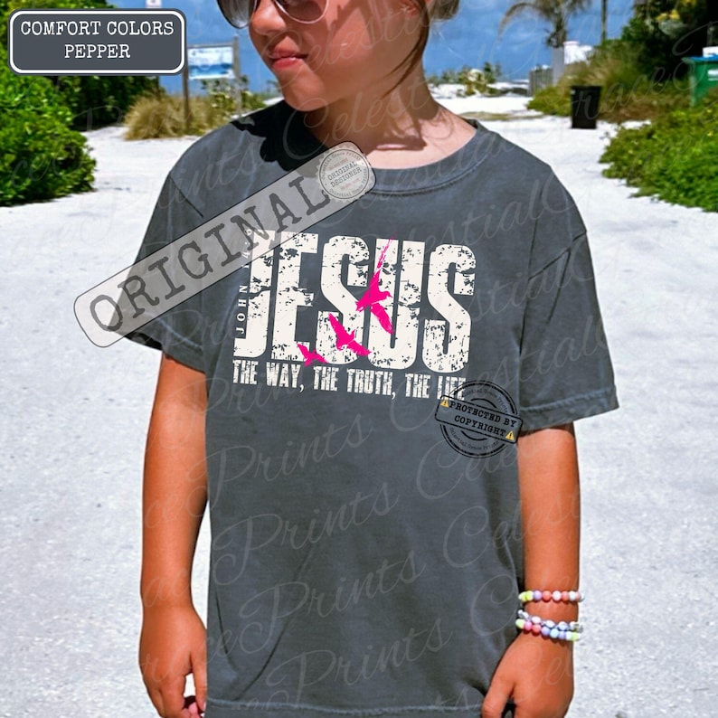 May include: A dark grey t-shirt with the word "JESUS" in white, and pink bird graphics. The text "THE WAY, THE TRUTH, THE LIFE" is below. The shirt is labelled "COMFORT COLORS PEPPER" and has a copyright notice.