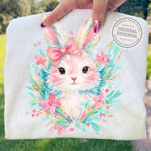 May include: White t-shirt featuring a cute bunny illustration. The bunny has pink fur, a pink bow, and is surrounded by pink flowers and teal accents. The design includes gold glitter details and the text "ORIGINAL DESIGNER".