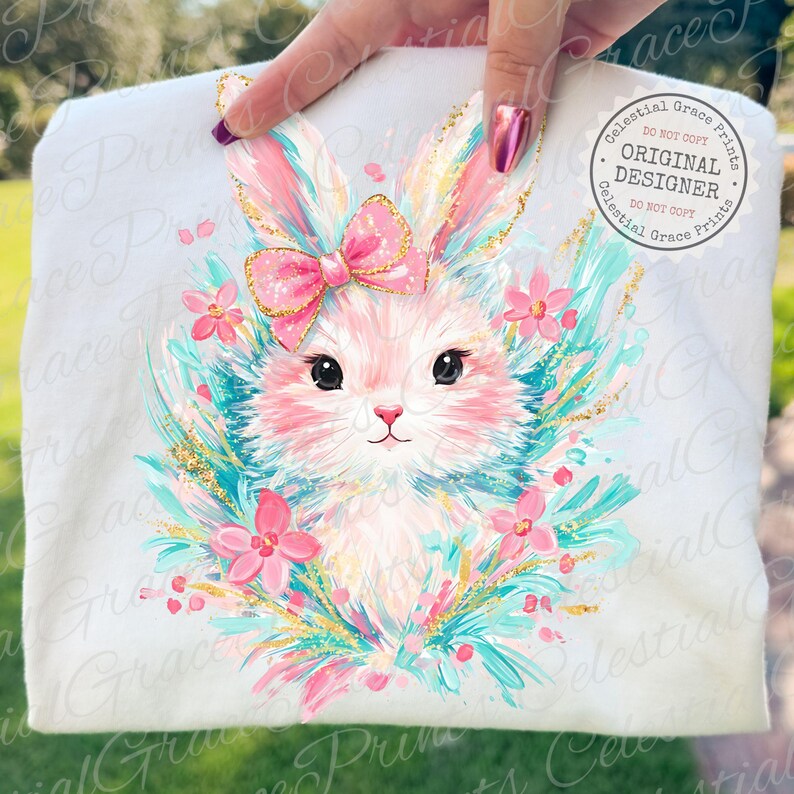 May include: White t-shirt featuring a cute bunny illustration. The bunny has pink fur, a pink bow, and is surrounded by pink flowers and teal accents. The design includes gold glitter details and the text "ORIGINAL DESIGNER".