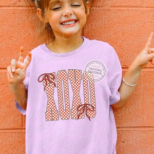 May include: A light purple t-shirt featuring the word "XOXO" in a red and white checkered pattern with red bows. The shirt has a round neckline and short sleeves. A circular graphic with text is on the upper right.