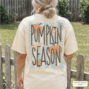 May include: An ivory short-sleeve t-shirt featuring a back graphic with the text PUMPKIN SEASON in dark brown letters. The design includes orange pumpkin illustrations, light blue vertical stripes, and small yellow dots on a light blue background.