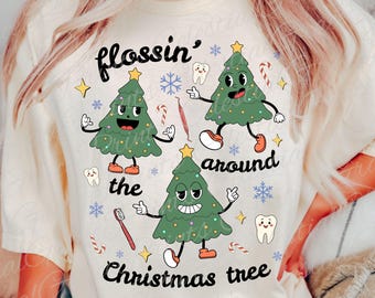 以下が含まれることがあります： クリーム色のTシャツ。3つの漫画のクリスマスツリーが踊って笑顔の楽しいグラフィックが特徴です。「flossin' around the Christmas tree」というテキストと、歯、歯ブラシ、雪の結晶などの歯科をテーマにしたイラストが描かれています。