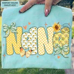 May include: A light blue t-shirt with the word "NANA" in large, decorative letters. The letters feature yellow polka dots, sunflower patterns, and gingham. A butterfly and bows add to the design. The shirt is labeled "Comfort Colors Chalky Mint."