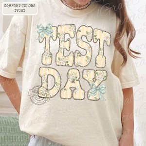 Peut inclure: T-shirt ivoire avec l'inscription "TEST DAY" dans une police fantaisie. Les lettres pr&eacute;sentent un motif &agrave; rayures jaunes et blanches, des motifs floraux et un contour l&eacute;opard. Des n&oelig;uds bleu clair ajoutent une touche d&eacute;corative.