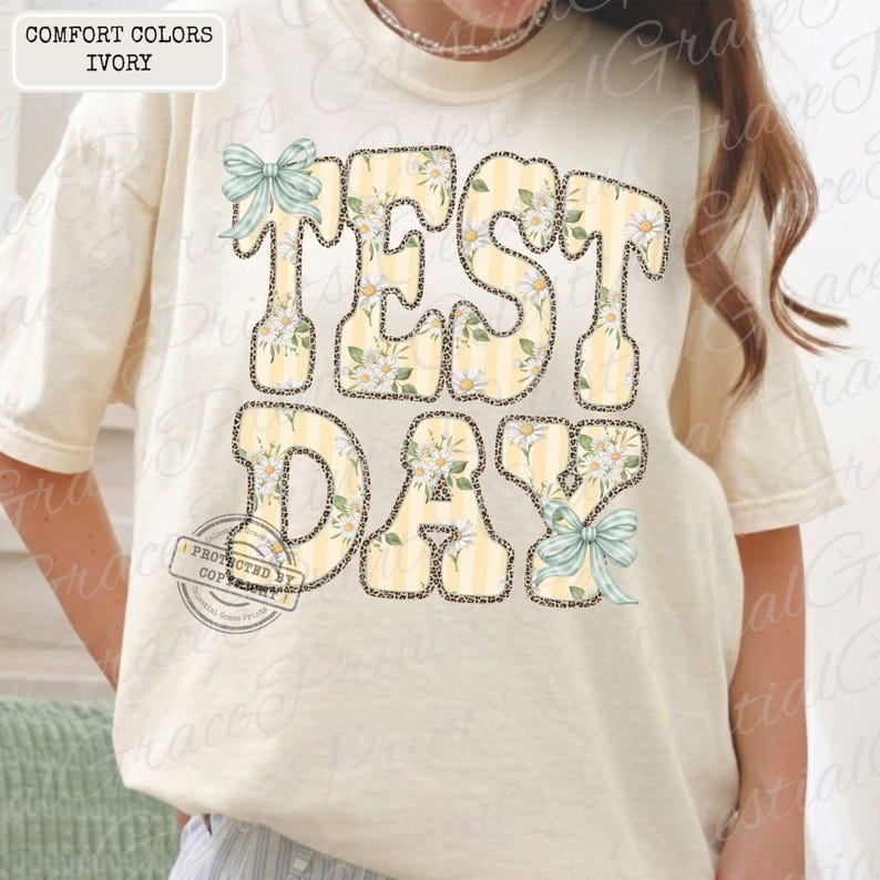 Peut inclure: T-shirt ivoire avec l'inscription "TEST DAY" dans une police fantaisie. Les lettres pr&eacute;sentent un motif &agrave; rayures jaunes et blanches, des motifs floraux et un contour l&eacute;opard. Des n&oelig;uds bleu clair ajoutent une touche d&eacute;corative.