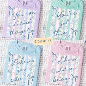 May include: Four pastel-colored t-shirts with inspirational messages. Each shirt features a unique design with text, ribbons, and floral accents. The messages include "You can do hard things," "You've got this," "Believe you can," and "Show what you know."