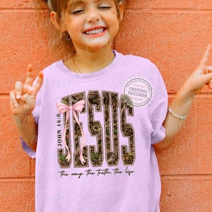 May include: A light purple t-shirt featuring the word "JESUS" in a camouflage pattern with a pink bow. The text "JOHN 14:6" is on the left side, and "The way. The truth. the life" is at the bottom.