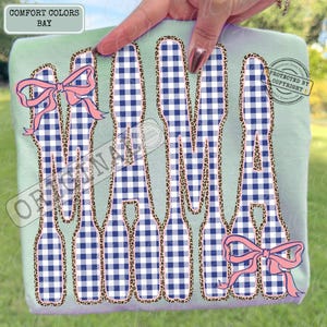 May include: A light green Comfort Colors sweatshirt featuring the word "MAMA" in a navy and white gingham pattern, outlined with leopard print. Pink bows are included in the design.