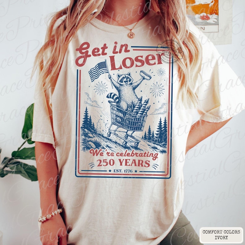 May include: Ivory-colored t-shirt with a graphic featuring a raccoon pushing a shopping cart filled with fireworks. The raccoon holds an American flag and a hot dog. Text reads "Get in Loser" and "We're celebrating 250 YEARS EST. 1776."