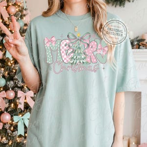 以下が含まれることがあります： 「MERRY Christmas」の文字がデザインされた、明るい緑色のTシャツ。文字はピンクと緑の模様、リボン、クリスマスツリーで装飾されています。快適でカジュアルなスタイルです。