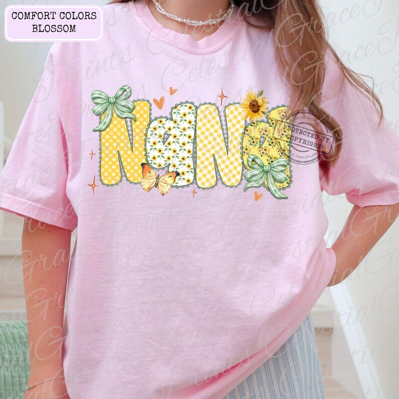 May include: A light pink t-shirt with the word "Nana" in a whimsical design. The letters are yellow with sunflower and gingham patterns, accented with green bows and butterflies. The shirt also has the text "Comfort Colors Blossom".