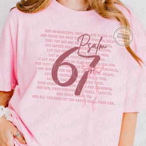 May include: A light pink t-shirt featuring the text "Psalm 67" in a large, stylized font, with a pink bow. The shirt also has a printed passage of text. A circular "Original Designer" sticker is visible.