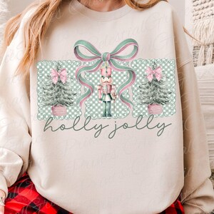 May include: A cream-coloured sweatshirt with a festive Christmas design. The design features a nutcracker, Christmas trees, and a large bow in pink and green, set against a checkered pattern. The words "holly jolly" are written in a script font.