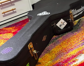 Könnte beinhalten: Schwarzer Gitarrenkoffer mit goldfarbenen Verschlüssen und Griff. Der Koffer hat mehrere Aufkleber, darunter einen mit der Aufschrift "Shure" in grünen Buchstaben. Der Koffer liegt auf einem bunten Teppich.