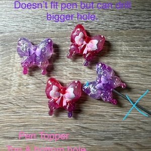 May include: Four butterfly-shaped pen toppers in shades of pink and purple. The toppers have a bow design and are translucent. Text on the image reads "Doesn't fit pen but can drill bigger hole" and "Pen topper Top & bottom hole".