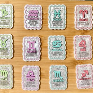 May include: A collection of twelve rectangular zodiac sign stickers. Each sticker features a unique zodiac symbol in various colors, with a decorative border and text describing the sign's personality. The stickers are arranged on a wooden surface.