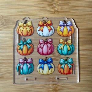 May include: A clear acrylic display stand featuring a grid of nine illustrated pumpkins, each adorned with a colorful bow. The pumpkins are arranged in three rows and three columns, showcasing various colors including orange, white, yellow, pink, and teal.