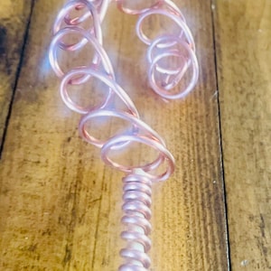 Puede incluir: Una escultura de alambre de cobre con una base en espiral y un diseño abstracto en espiral en la parte superior. La escultura es de color rosa claro y se fotografía sobre un fondo de madera.