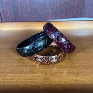 May include: Three stacked bangle bracelets made of snakeskin. The top bracelet is purple, the middle bracelet is black, and the bottom bracelet is brown.