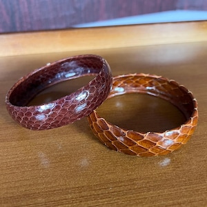 May include: Two leather bracelets with a snakeskin pattern. One is brown and the other is a darker reddish brown.
