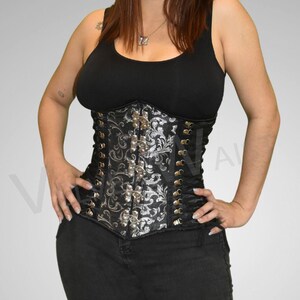 May include: A black and silver corset with a floral pattern and gold-colored accents. The corset has multiple hook-and-eye closures down the front and is worn with a black tank top and black jeans.