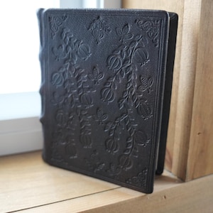 May include: A black, embossed leather journal with floral and butterfly designs. The cover features a detailed pattern of leaves, flowers, and butterflies. The journal is standing upright on a wooden surface, near a window.