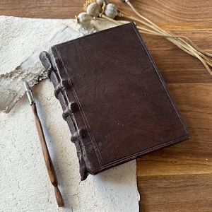 May include: A dark brown leather-bound journal with a simple design, resting on a sheet of handmade paper with a wooden pen.