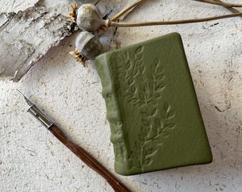 Pode incluir: Um pequeno diário de couro verde com um design floral em relevo na capa. Uma pena de mergulho de madeira está sobre uma superfície branca texturizada ao lado do diário.