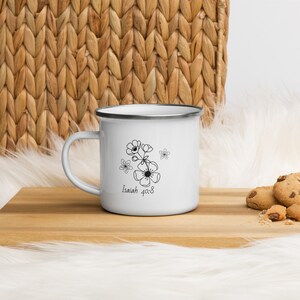 Puede incluir: Taza de esmalte blanco con un diseño floral negro y el texto "Isaías 40:8". La taza tiene un borde y un asa plateados. La taza está sobre una superficie de madera.