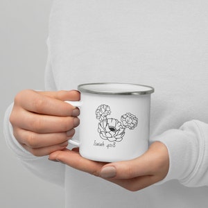 Puede incluir: Taza de esmalte blanco con borde y asa plateados. La taza presenta un diseño floral negro con el texto "Isaías 40:8". La taza está sujeta por una persona.