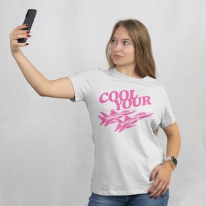 Może przedstawiać: Biała koszulka z różowym napisem "COOL YOUR" i dwiema różowymi grafikami odrzutowców. Koszulka wykonana jest z miękkiego materiału.