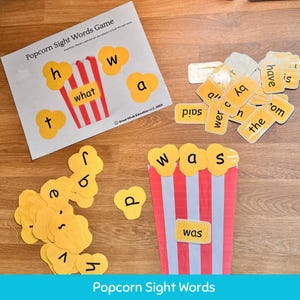 May include: A children's educational game with a popcorn theme. The game includes a sheet with a popcorn bucket and sight words, along with individual popcorn-shaped cards with letters and words. The words "what" and "was" are visible.