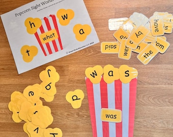 May include: A children's educational game with a popcorn theme. The game includes a sheet with a popcorn bucket and sight words, along with individual popcorn-shaped cards with letters and words. The words "what" and "was" are visible.