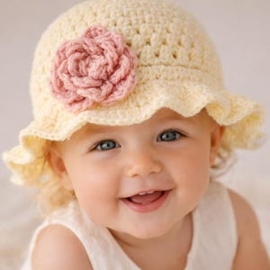 May include: A cream-coloured crochet hat with a pink flower. The hat has a frilled brim. The person in the image is wearing a white dress and smiling. The background is a soft, neutral colour.