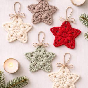 May include: Five star-shaped ornaments made of crocheted yarn in cream, dusty rose, red, and sage green. Each ornament features a central puff stitch pattern and a small jute twine loop for hanging, tied with a decorative bow at the top.