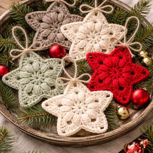 May include: A collection of five star-shaped crochet ornaments in cream, red, sage green, and taupe. Each star features a textured puff stitch center and a jute twine loop for hanging. The ornaments are arranged on a round metal tray with pine branches.