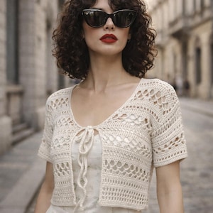 Puede incluir: Rebeca de crochet color crema, de manga corta, con cierre de lazo. El diseño calado presenta un patrón repetitivo. La rebeca se lleva sobre un vestido de color claro. La persona lleva gafas de sol negras y pintalabios rojo.
