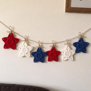 May include: A garland featuring six crocheted stars in red, white, and blue, suspended from a thin natural twine string. Each star is individually tied to the twine with a small bow, alternating in color pattern across the length of the string.