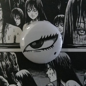 May include: A white button with a black and white eye design, set against a black and white manga illustration background. The button features a detailed eye with long eyelashes. The background shows multiple manga figures with long black hair.