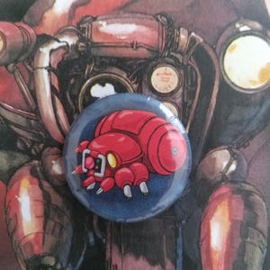 Può includere: Una spilla rotonda con finitura lucida che presenta un'illustrazione grafica di una piccola creatura robotica rossa a più zampe, con una piastra frontale bianca e un occhio giallo, su uno sfondo circolare blu scuro.