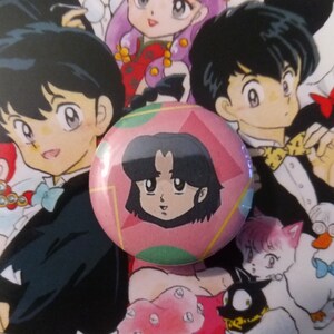 May include: A pink and green button with a cartoon image of a girl with black hair and a pink shirt. The girl is looking to the right.
