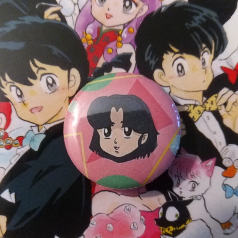 May include: A pink and green button with a cartoon image of a girl with black hair and a pink shirt. The girl is looking to the right.