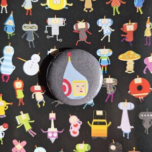 Può includere: Un bottone rotondo grigio scuro con un design di personaggi dei cartoni animati. Il bottone presenta una forma a goccia azzurra con una mezzaluna rossa e una faccia quadrata gialla. Lo sfondo è nero con vari adesivi di personaggi dei cartoni animati.