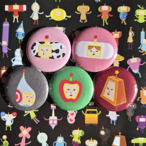 Può includere: Una collezione di cinque bottoni rotondi e colorati con facce di cartoni animati. I bottoni presentano sfondi rosa, verdi, grigi e marroni con vari disegni, tra cui una mela e un motivo a mucca. Lo sfondo è nero con personaggi dei cartoni animati.