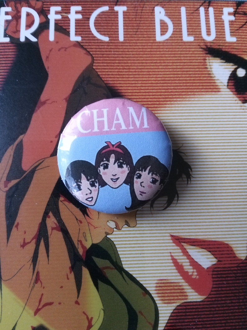 May include: A blue and pink button with the word "CHAM" printed on it. The button features a cartoon image of three women with black hair.