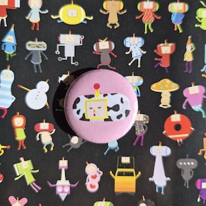 May include: A pink button with a cartoon robot design, featuring a yellow frame and black and white spots. The button is surrounded by various colorful cartoon character stickers on a black background.